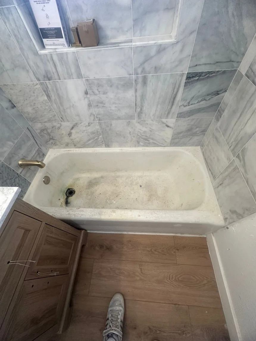 A bathtub with heavy gray staining across the base, set in a bathroom tiled in large veined marble-effect tile.